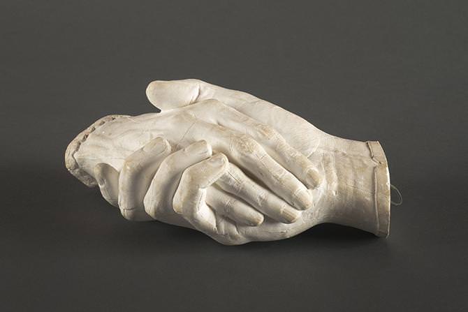 Sculpture. Plaster cast of two right hands holding each other. The hands cut off at the wrists, and one is edged in a straight cuff and the other in scalloped cuff. In this view, one rests along the palm of the other.